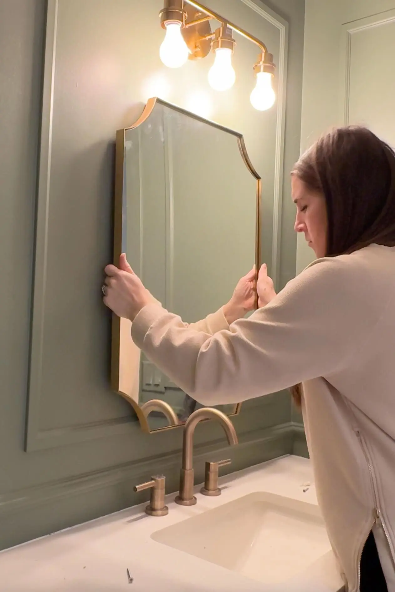 Woman in a cream sweater carefully adjusting a brass-framed mirror above a white sink with brass fixtures, set against a dark green bathroom wall and illuminated by a brass light fixture with three exposed bulbs.